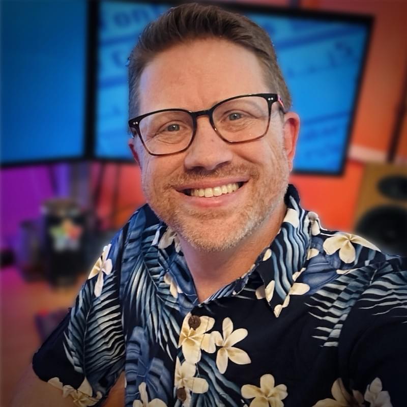 Joe Workman, smiling in a floral shirt in front of two monitors