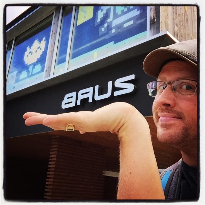 Joe in a cap and glasses, palm held up under the ZURB sign on the office building, with pixel-art space invaders in the window above