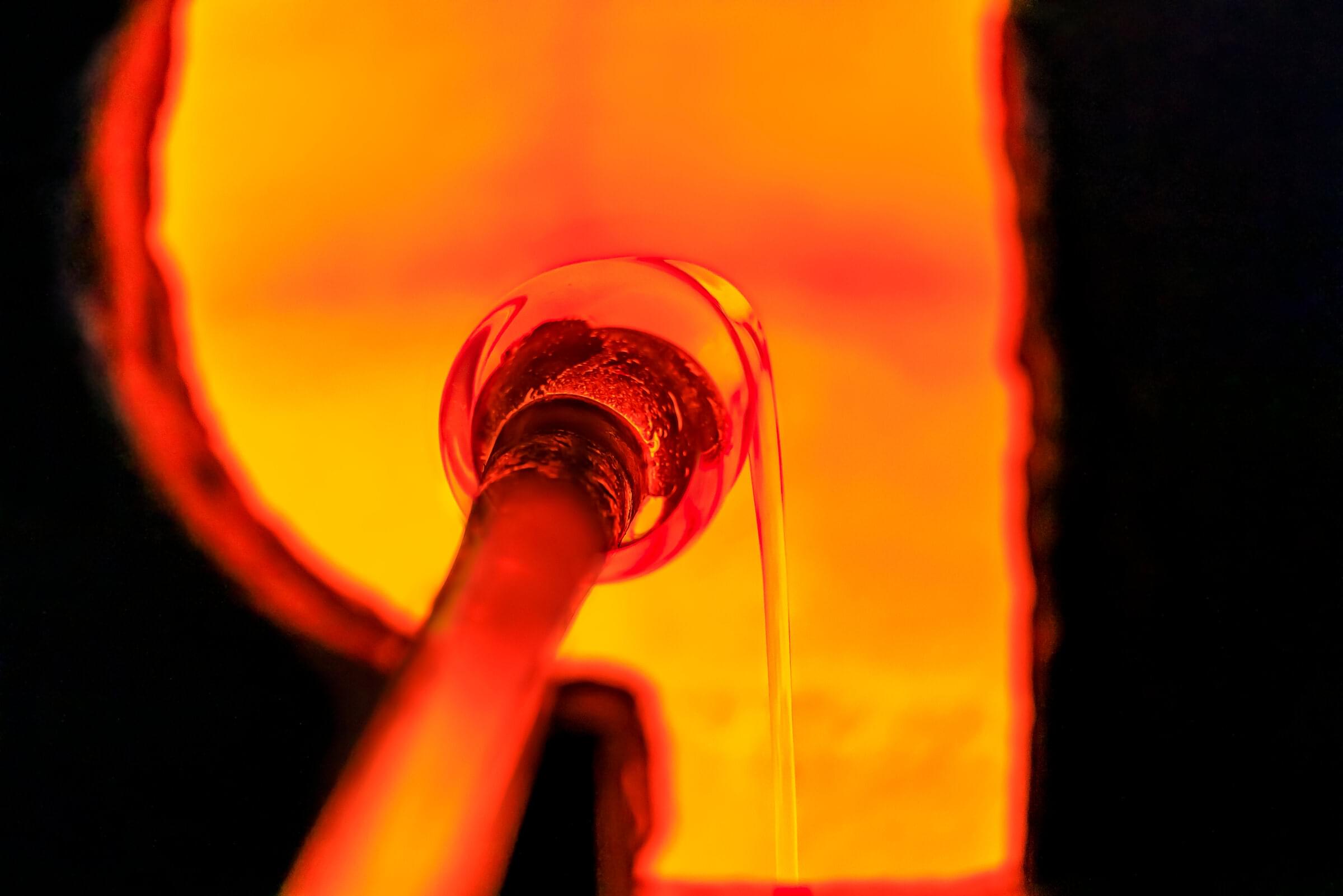 A gather of molten glass turning on the end of a blowpipe inside the glory hole, a thread of it falling away, all lit orange by the fire.