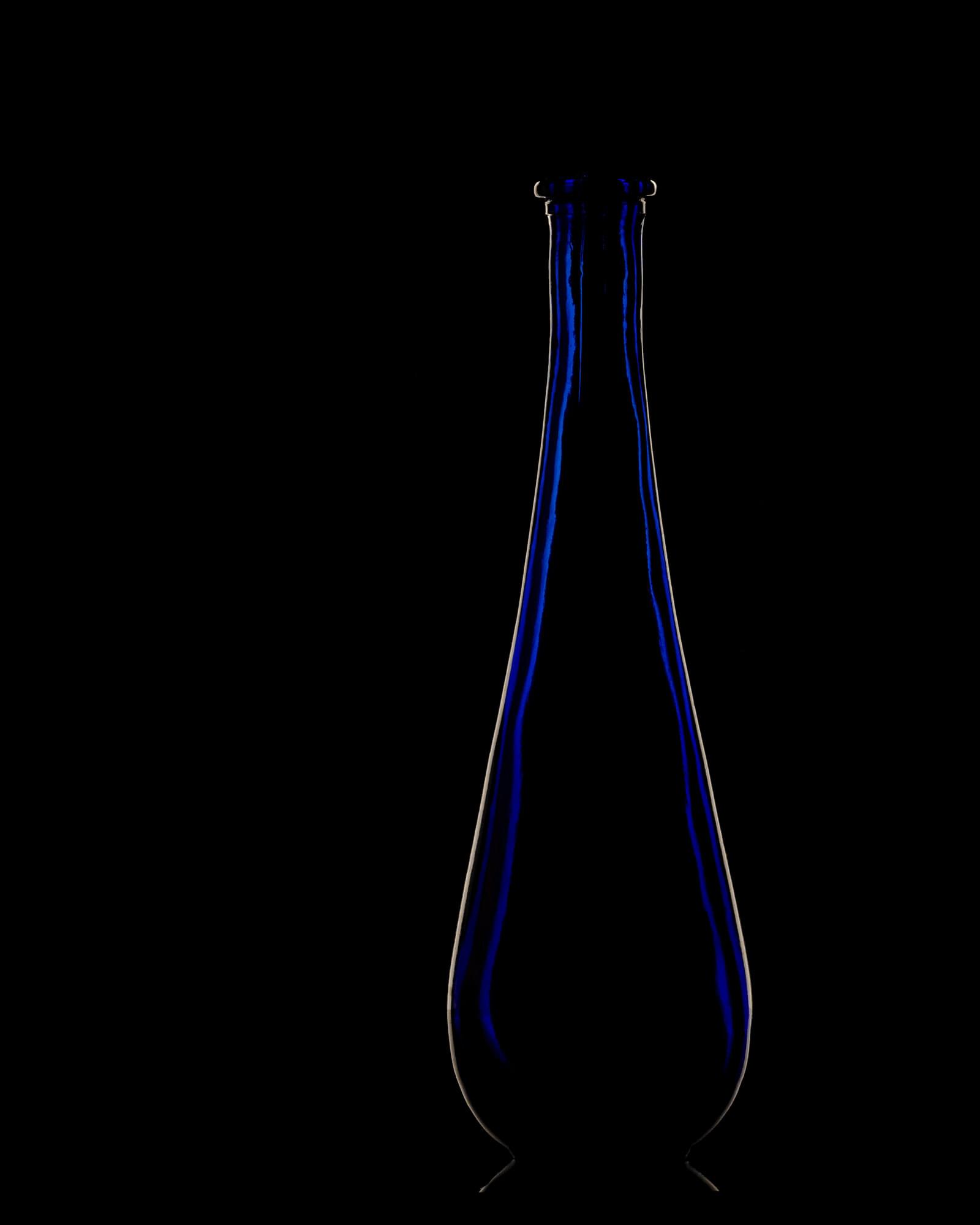 A tall teardrop bottle of cobalt glass in the dark, lit from behind so only its edges and the blue in its walls show.