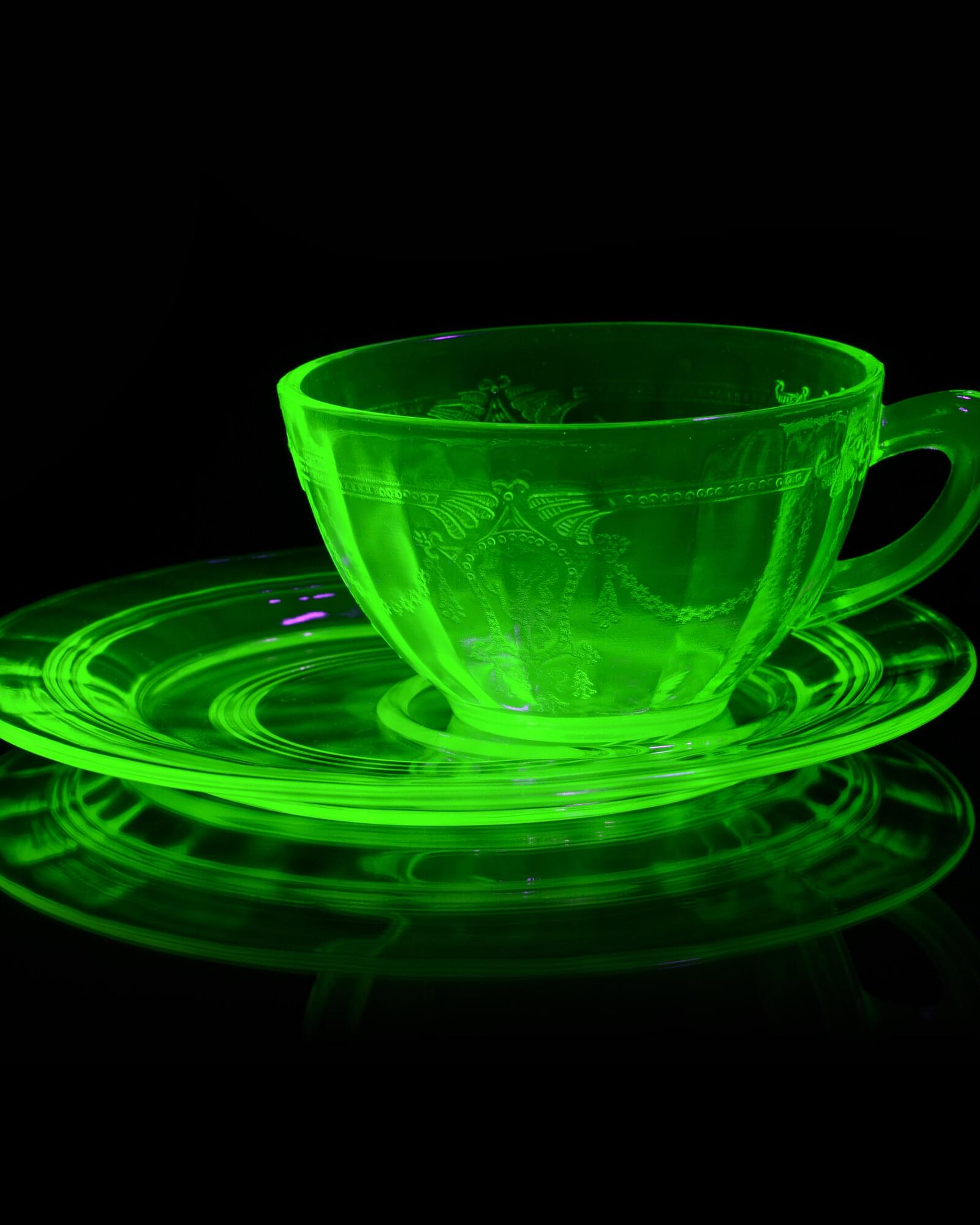 Cup and Saucer: a uranium glass cup and saucer glowing bright green under ultraviolet light, doubled in a black mirror.