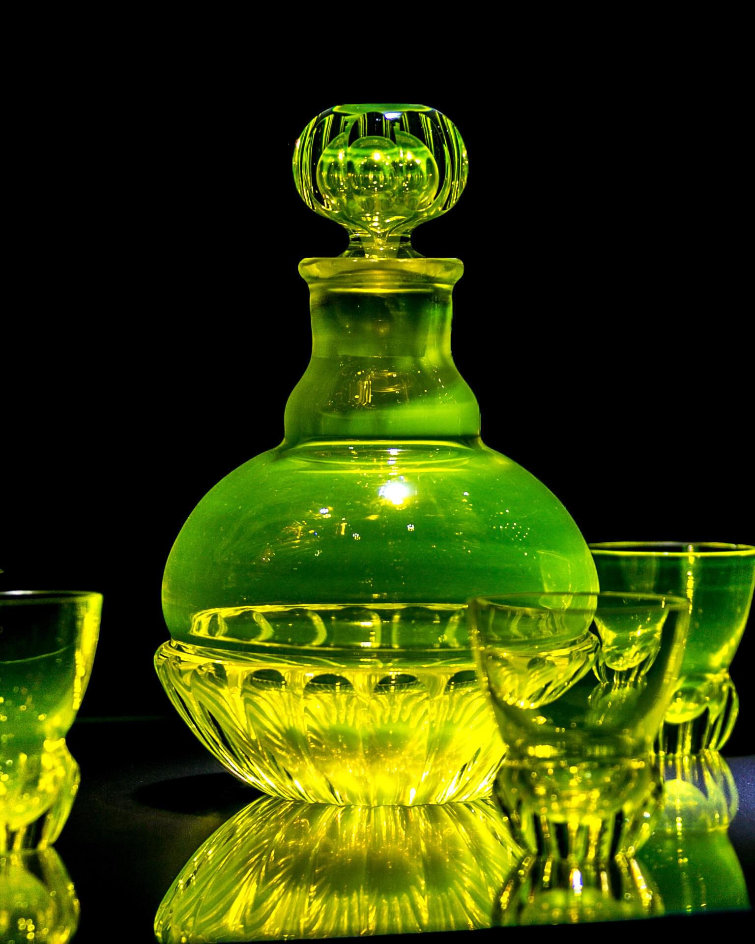 Service for Six: a stoppered decanter and small cut glasses glowing yellow-green under ultraviolet light in the dark.