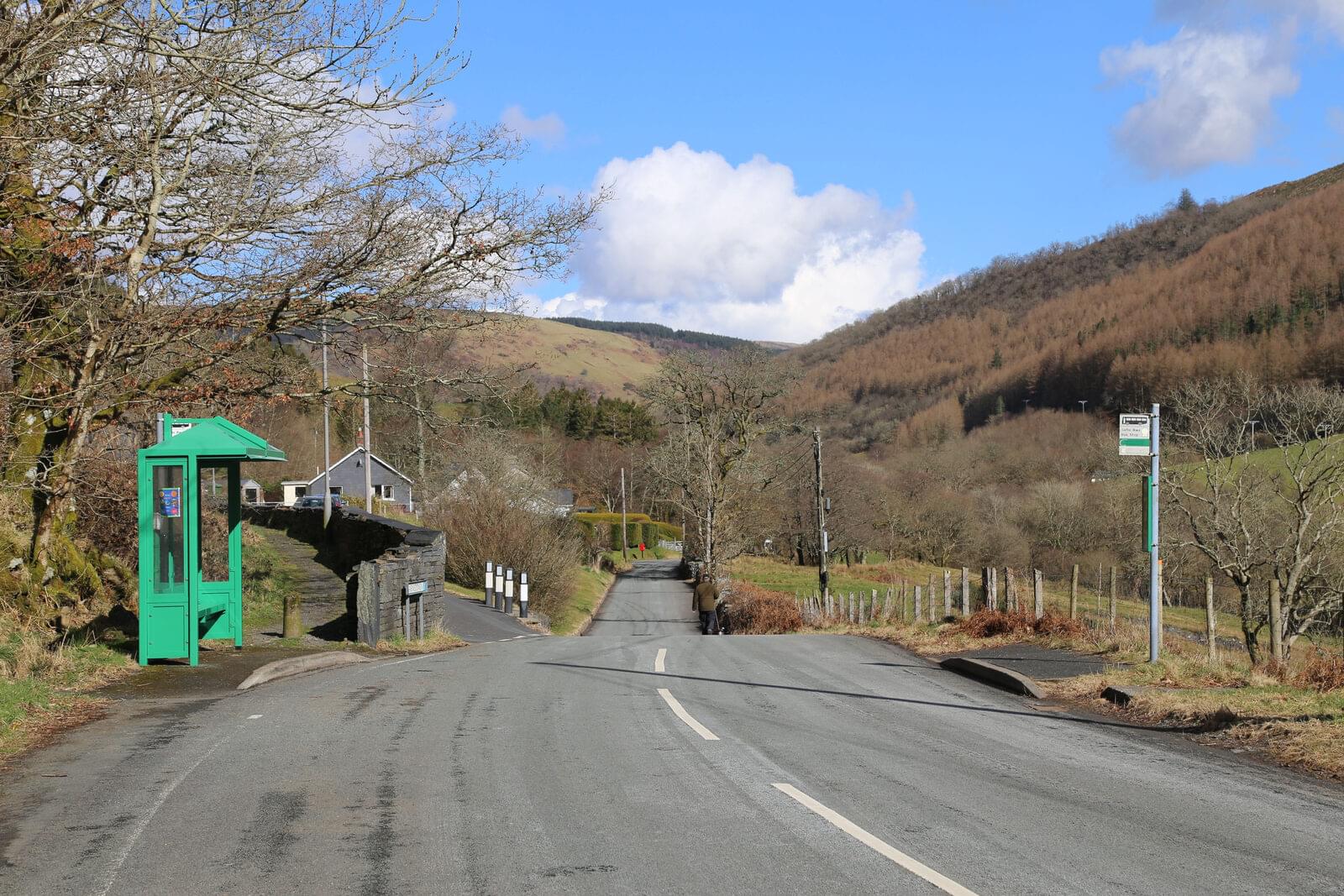 A green bus shelter beside a quiet valley road, wooded hills behind