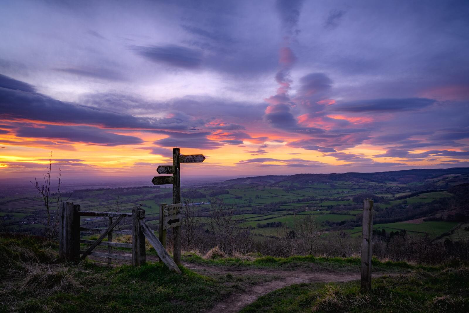 A fingerpost and gate on a hilltop path at sunset, the vale spread out below under a streaked sky